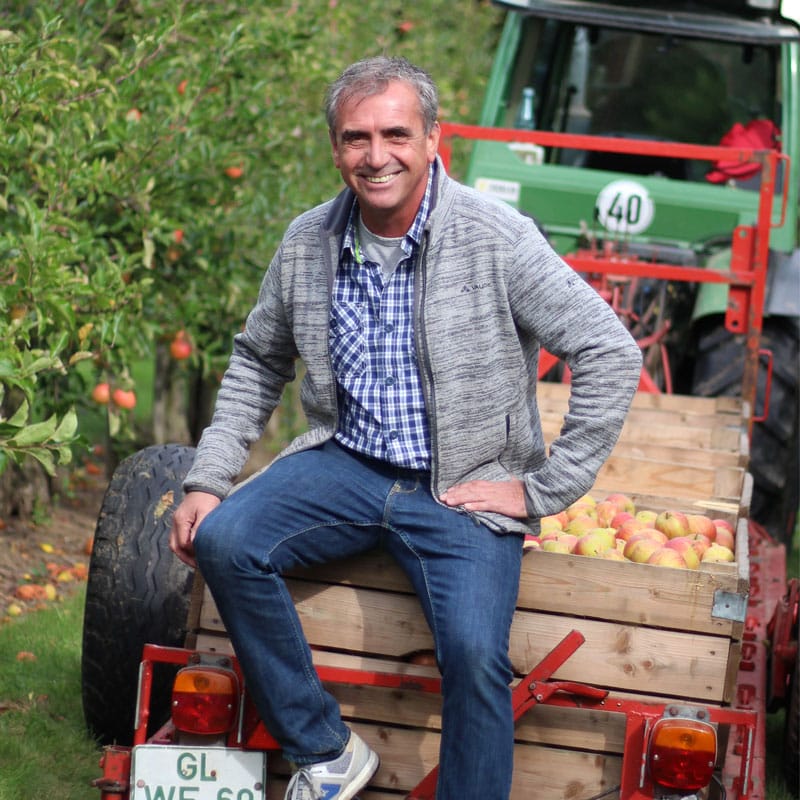 Michael Altmeyer sitzt auf eine Apfelkiste die am Trecker angebracht ist, inmitten seinen Apfelbäumen.
